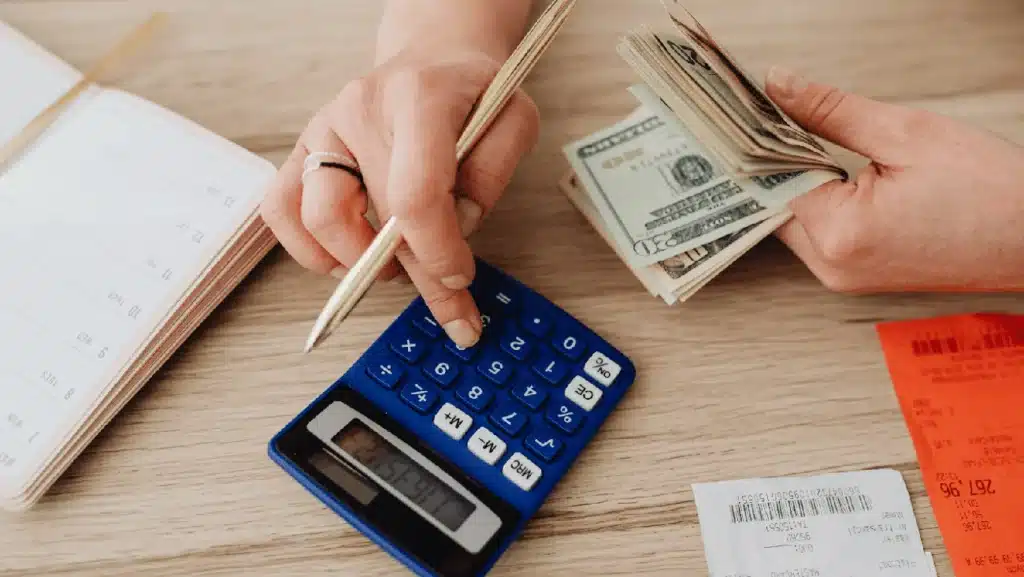 Person using a blue calculator and holding cash with receipts and a planner on a wooden table.